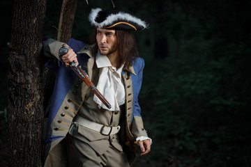 Fototapeta na wymiar 18th-century officer with flintlock gun stands by tree in forest