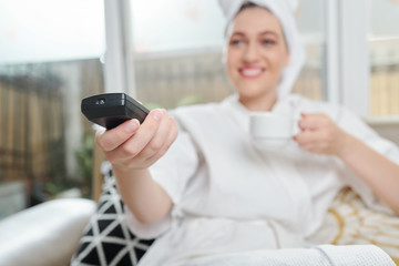 Fototapeta na wymiar Pretty smiling young woman drinking cup of tea and checking channels on her tv, selective focus
