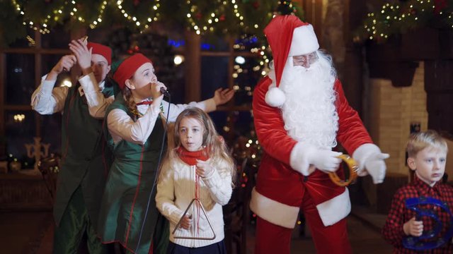 Festive atmosphere in Santa's residence. Santa Claus, two elves and children dancing and singing together at Christmas.