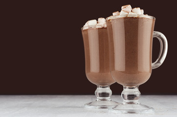 Holiday chocolate drink in elegant transparent goblet with marshmallows, cocoa powder on dark brown background, white wood board, copy space.