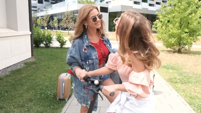 Two girls are staning with a scooter on a pathway. They wear sunglasses. There is a building on a backgroung.