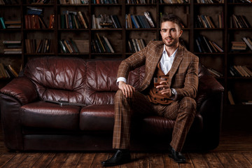 young successful man in a brown kosmtyum drinks whiskey. bearded businessman sitting in a library...