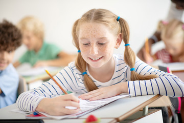 Smiling girl with freckles smiling while writing at the lesson