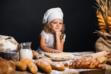 Charming little girl with curly hair in white apron and hat standing at table kneading bread dough...