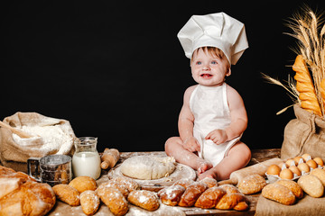 Charming toddler baby in hat of cook and apron sitting on table with bread loaves and cooking ingredients laughing happily