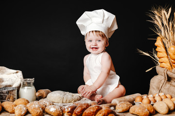 Charming toddler baby in hat of cook and apron sitting on table with bread loaves and cooking ingredients laughing happily