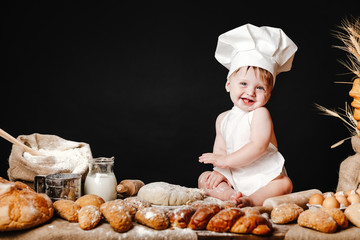 Charming toddler baby in hat of cook and apron sitting on table with bread loaves and cooking ingredients laughing happily