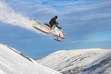 a snowmobile flies in the mountains with a large snow trail against a bright blue sky with clouds. Snowmobile jump at high altitude. bright snowmobile and looking at the camera. super quality photo