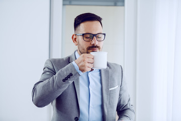Attractive caucasian unshaven man in suit and with eyeglasses standing next to window and smelling fresh coffee.