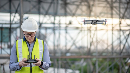 Obraz premium Asian engineer man flying drone over construction site. Male worker using unmanned aerial vehicle (UAV) for land and building site survey in civil engineering project.