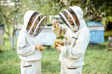 Two beekepers in protective uniform standing together with honey in the jar, tasting fresh product...