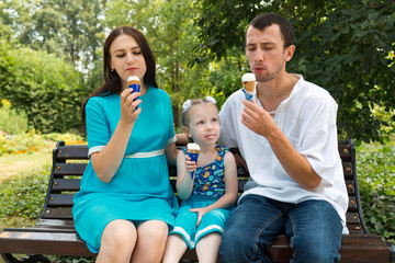 Father, mother and daughter sit on bench and eat ice cream. Woman pregnant.