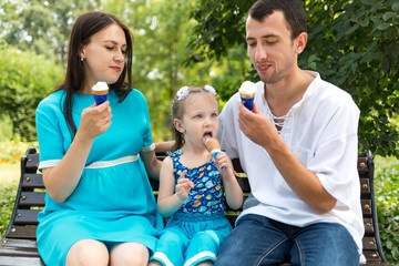 Fototapeta premium Father, mother and daughter sit on bench and eat ice cream. Woman pregnant.
