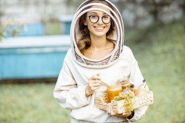 Portrait of a cheerful female beekeeper in protective uniform standing with fresh honey and sweet...