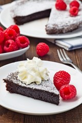 Piece of chocolate sticky brownie cake, swedish dessert Kladdkaka, on plate, garnished with icing sugar, whipping cream and raspberry, vertical, closeup