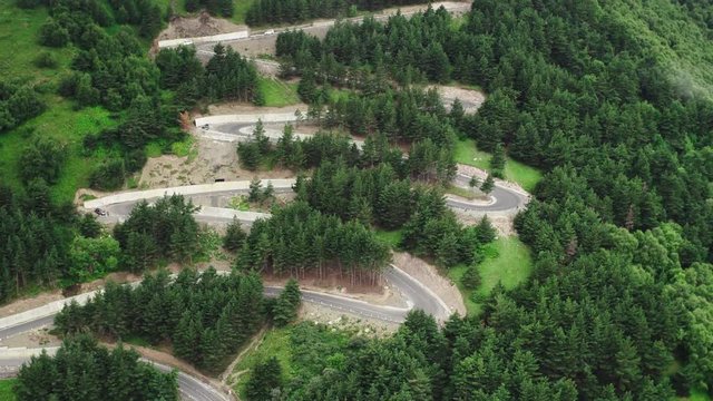 Cars driving on serpentine winding road through lush dense spruce forest on mountain slope. Curvy switchback highway with hairpin turns snaking through the woods - Aerial drone view