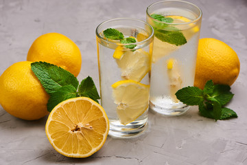 Glasses filled with pure water with lemon slices surrounded by a few whole lemons, fresh mint leaves on the gray concrete background. Close up