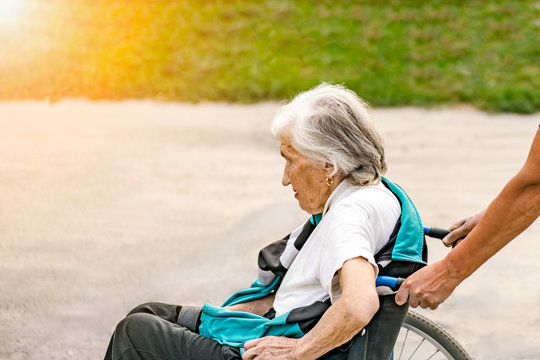 Old paralyzed grey-haired woman on a disable carriage moving on the street with relative. Woman after stroke who cannot move by herself