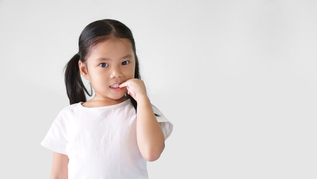 Asian child cute or kid girl nail biting on finger and smiling with anxious or thinking enjoy on preschool or 5 years old and wear white T-shirt and white background isolated with copy space