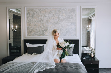 Smiling blonde bride in a white coat and lingerie, with a long veil sits on a sofa with a bouquet....