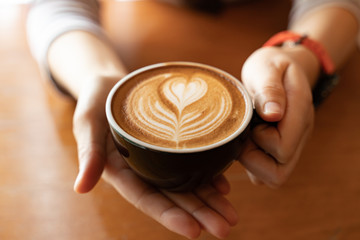 Close up Cup of coffee latte in coffee shop.Female hands holding a cup of coffee cup with heart shaped latte art foam on black wood table.