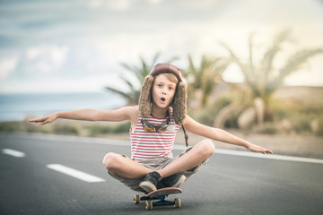 Fototapeta na wymiar Young blond boy plays aviator sitting cross-legged on skateboard with arms outstretched to fly smiling child imitates plane flying on airport runway. Concept image of a plane taking off for a vacation