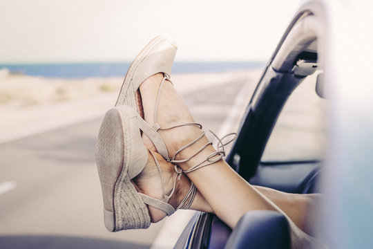 Women's feet out of the car window with a road and the sea in background. Crossed legs of young girl with trendy shoes. Female relaxing during summer adventure. Freedom, travel, female's fun concept.