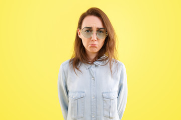 Offended stylish young woman in a blue shirt and glasses made a sad face and looks in a camera on a yellow background. Advertising space