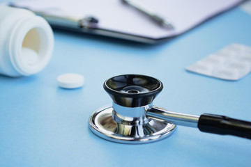Medical stethoscope on a light blue background. In the background are pills, blank clipboard and pen. Healthcare and medical concept. 