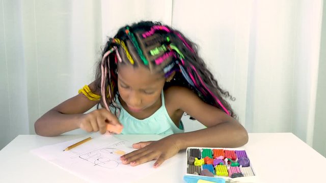 Jamaican mixed ethnicity girl with colored braids sitting at the table. The child coloring a picture with orange chalk, creative activity