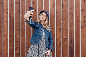 Young beautiful girl student stands at wooden background and makes selfie on the smartphone for friends. Gadgets and people. Technology in the Modern World, Narcissism and Ego