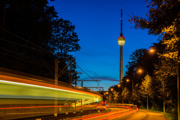 Fototapeta na wymiar Germany, Fast motion of driving train alongside traffic road with illuminated tv tower building behind in magical twilight mood after sunset in summer