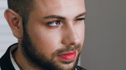 Obraz premium Portrait of young handsome man with makeup on his face winks to somebody and smiles. Portrait of bearded metrosexual or gay man blinks