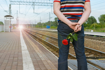 view of the man behind, who holds a red rose flower, to stand on the platform of the railway station against the background of headlights approaching from afar train