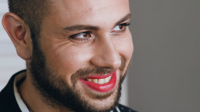 Portrait of young handsome man with makeup on his face winks to somebody and smiles. Portrait of bearded metrosexual or gay man with toothy smiling