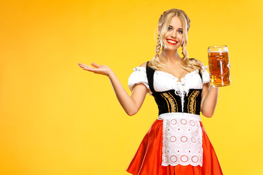 Young sexy Oktoberfest girl waitress, wearing a traditional Bavarian or german dirndl, holding big beer mug with drink isolated on yellow background. Woman pointing to looking left. - Powered by Adobe