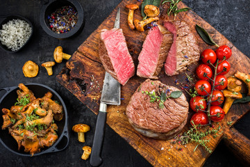 Gebratenes dry aged Rinderfilet Medallion Steak natur mit Pfifferlingen und Tomaten als Draufsicht auf einem Holz Schneidebrett 