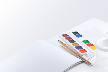 Side view of beautiful watercolor paints, brushes and an open drawing album with blank pages on a white background with empty space for text