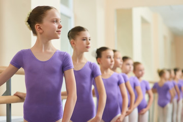 Happy young ballerina at dance lesson. Group of young ballet girls standing in row at ballet barre...