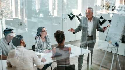 Discussing new project . Mature businessman standing near whiteboard and pointing it while his colleagues sitting at the office table and listening to him