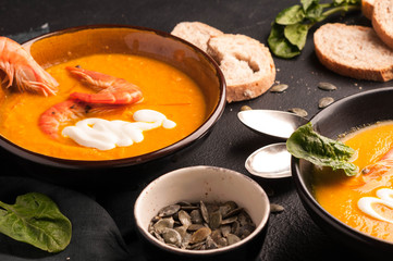 side view of pumpkin soup with prawns and sour in bowls near homemade bread, seeds, herbs on black background