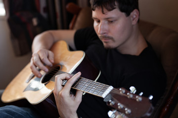 Obraz premium close-up portrait of a young man in black t-shirt playing an acoustic guitar. Focus is on his hand holding a chord string for a chord in interior