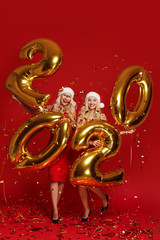 Happy New Year. Women tweens With Balloons Celebrating At Christmas Party. Portrait Of Beautiful Smiling Girls In fashion Dresses Throwing Confetti, Having Fun With Gold 2020 Balloons On red Backgroun