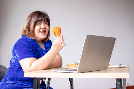 Chuby woman eating a large piece of fried chicken in the living room with a lot of fast food on the table. She stuck her tongue out of hunger and thirst and stared at the object with excitement.