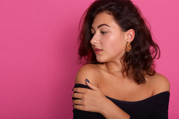 Obraz premium Horizontal shot of young beautiful woman looking aside, posing isolated over rosy background with bared shoulders, wearing black shirt, having wavy hair. Copy space for advertisment or promotion text.