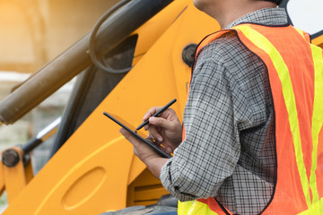 Engineering wearing a white safety helmet standing In front of the backhoe And are using tablet for check the blueprint with construction