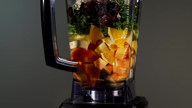 Making fresh fruits cocktail shake. Papaya, banana slices, curly kale leaves and sprouts in blender ready to be mixed, machine rotating on grey. Healthy food, dieting