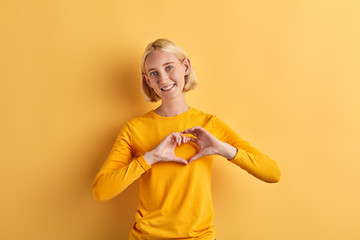 positive charming beautiful woman makes heart gesture with fingers, isolated yellow background, studio shot, positive feeling and emotion , I love you