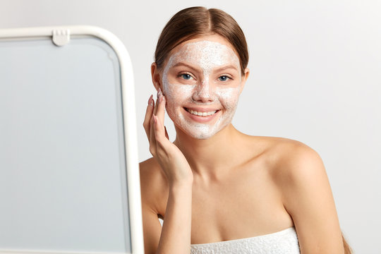 cheerful young woman standing in front of the the mirror with mask on her face. girl applying facial cosmetic mask after taking a shower in bathroom.isolated white background, studio shot