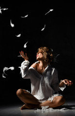 beautiful young blonde is sitting on the floor in one white shirt having fun with feathers. Stormy night.